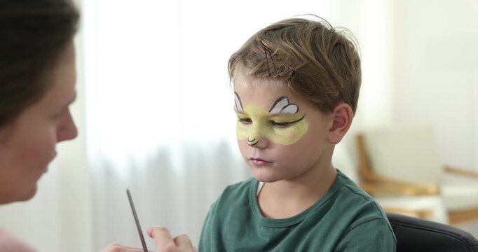 Woman drawing bee on boy's face indoors, closeup