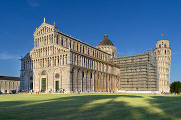 Iconic Pisa complex featuring the cathedral, Baptistery, and Leaning Tower in a sunlit Piazza dei Miracoli. Historic, architectural marvel with tourists exploring the grass lawn under a clear blue sky