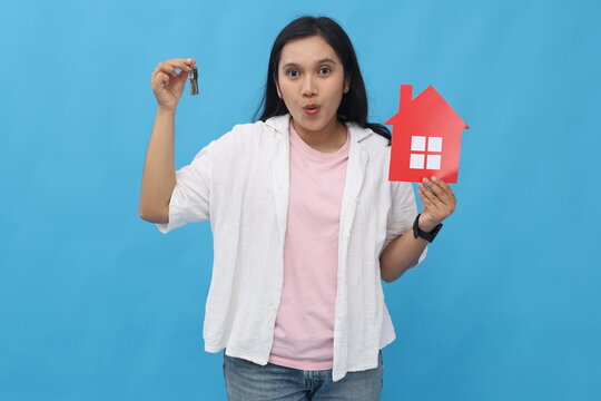 Female hands holding a small red paper house image and house keys, posing on a blue background wall in a studio with copy space for promotional or design content, real estate and family home concept. - Powered by Adobe