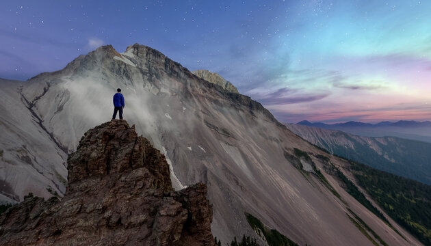Person Standing On Rocky Peak Under Aurora Borealis Over Mountain Range at Dusk