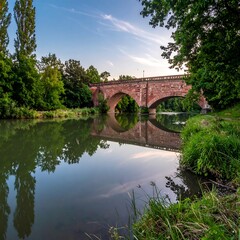Fototapeta premium Stone bridge over a calm river with lush greenery and bright sky