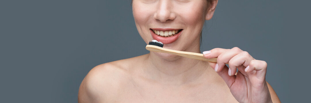 Smiling caucasian female brushing teeth with eco-friendly bamboo toothbrush on grey background.