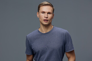 Obraz premium Concerned young man with short blond hair wearing casual blue t-shirt looking surprised or confused isolated on gray background studio portrait. People emotion concept