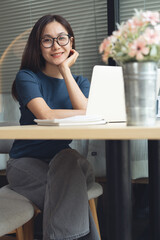 Asian business woman sitting at table, looking at the camera, with laptop computer on table at coffee shop, remote working, digital nomad, remote work concept, vertical composition