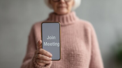 An elderly woman in a cozy sweater holds a smartphone displaying the message "Join Meeting," suggesting a virtual gathering.