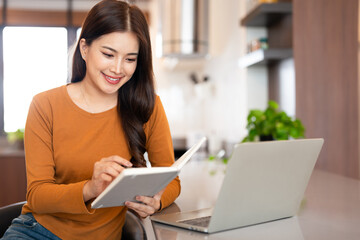 Joyful woman sitting at kitchen table and making notes in her notebook with pen.