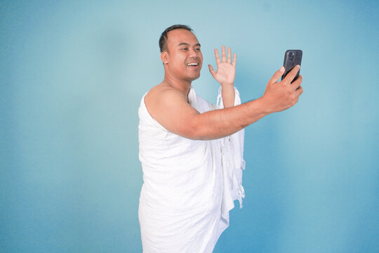 young Asian Muslim man wearing ihram doing video call pose while holding smartphone, isolated blue background. Hajj concept. islam - Powered by Adobe