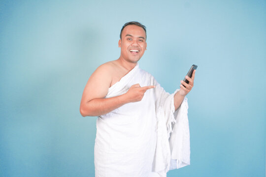 young Asian Muslim man wearing ihram doing video call pose while holding smartphone, isolated blue background. Hajj concept. islam