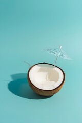 Minimal tropical composition with half a coconut filled with creamy drink and a small cocktail umbrella on a bright turquoise background, symbolizing summer relaxation, exotic vacation, and refreshmen
