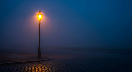 Solitary illuminated lamp post stands in the dark misty night by the water