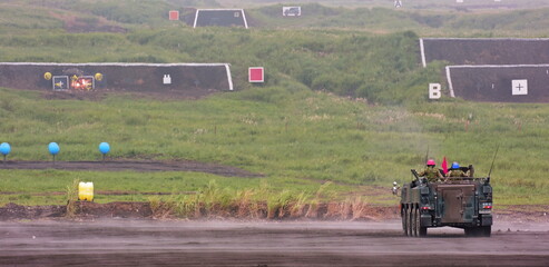 Soldiers aboard the military vehicle navigate a wet, dusty training area with multiple backstop targets showing signs of impact and a light haze lingering over the field.