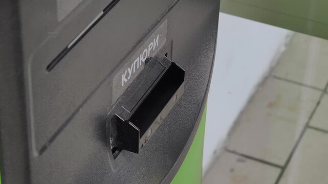 Side angle close-up person's hand in brown sleeve placing a Ukrainian Hryvnia banknote into the cash acceptor of a self-service kiosk, processing an electronic payment transaction in Ukraine.