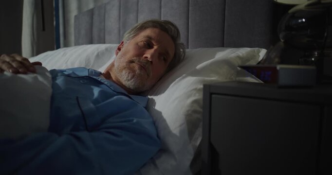 Middle Aged Man Wakes Up Early in Morning in Bedroom Filled With Sunlight. Gray Haired Man Stretches in Bed, Puts on Glasses. Concept of Concept Morning Routine, Relaxation, Comfort, Cozy Morning.