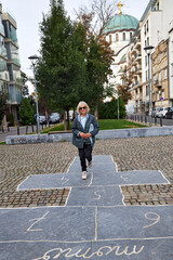 Elderly woman standing on a hopscotch game drawn with chalk on cobblestone pavement in a city street. Urban nostalgia and playful spirit meet in this cheerful moment of childhood memories and outdoor 