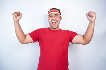 Naklejka premium A young Asian man with a happy successful expression, raised a fist wearing red shirt and flag headband isolated by white background. Indonesia's independence day concept.