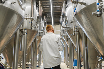 Man moving between rows of large stainless steel fermentation tanks in a modern beer production facility