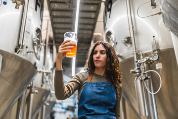 Woman brewer evaluating beer quality, holding a tasting glass, standing in a modern craft beer production facility