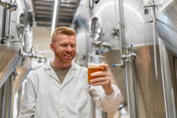 Smiling brewer examining beer in a glass for quality, standing in a small brewery with stainless steel tanks