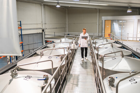 Man in lab coat walking along stainless steel fermentation tanks, holding a digital tablet and checking beer production data - Powered by Adobe