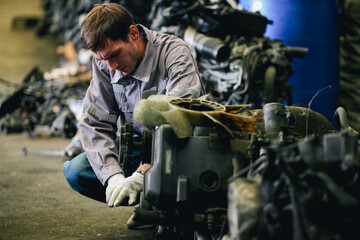 White man technician checking used car damaged engine block at scrap yard warehouse recycle area part. Maintenance engineer inspecting rust oily auto motor old spare part in junkyard for reuse service