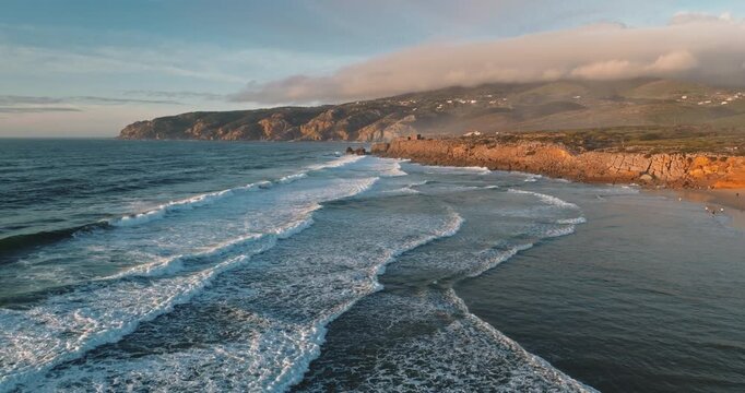 Portugal, Lisbon: Praia do Guincho coastline with white foam waves crashing on golden rocky cliffs under wide clear sky, remote coastal beach aerial landscape. Travel background. Drone flight footage
