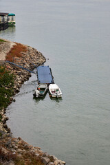 Two small boats moored at a blue floating dock on the rocky Danube River bank near Novi Sad, Serbia. Peaceful riverside view with greenery, autumn tones, and calm water under cloudy sky