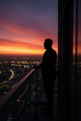 Fototapeta premium A silhouette of a man standing on a skyscraper balcony at sunset, with city lights below, a symbol of financial success.