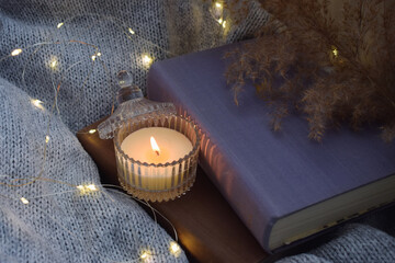 Lit candle in glass holder beside closed book surrounded by soft knitted blanket dried plants and...