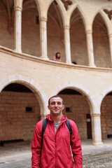 Young handsome man in red jacket inside of an old castle in Palma de Mallorca. Portrait of a young athletic man. Happy face expression. Travel mood. Explorer tourist. Travel agency advertisement. 