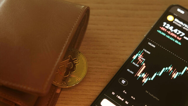 Bitcoin, finance, investment, and digital economy concept with physical coin beside wallet and smartphone displaying cryptocurrency trading chart