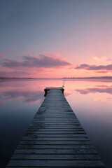 Fototapeta premium A serene view of a wooden pier extending into a calm lake reflecting a pastel pink sunrise. Evokes peace, solitude, and the beauty of nature. Ideal for travel, mindfulness, or lifestyle content.