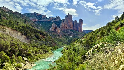Mallos de Riglos in Spain features striking, vertical rock formations set against a backdrop of clear blue sky and scattered clouds. The towering red and brown cliffs, surrounded by dense green pine  © Julija
