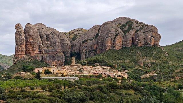 A picturesque view of the village of Aguero, Spain, nestled below the dramatic Mallos de Aguero rock formations. These imposing conglomerate cliffs tower over the quaint stone buildings of the village