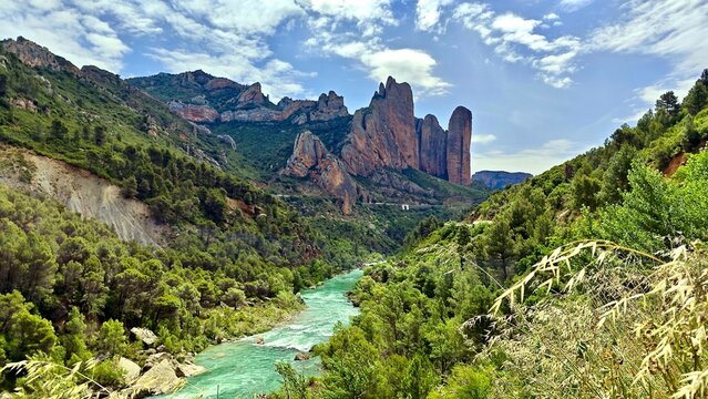 Mallos de Riglos in Spain features striking, vertical rock formations set against a backdrop of clear blue sky and scattered clouds. The towering red and brown cliffs, surrounded by dense green pine 