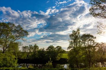 Majestic blue sky with dramatic clouds above a tranquil river framed by vibrant green trees and a soft sunset glow