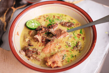 A top-down view of Soto Kuning Bogor, a traditional Indonesian yellow beef soup. The savory broth is garnished with a lime slice and fresh herbs, with a spoon resting in the bowl. Bogor popular Soup