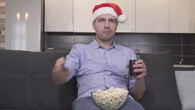 Video of serious concentrated guy in christmas hat eating popcorn, drinking sida and watching tv alone in room. Switching channels with remote control. Christmas time or new year period.