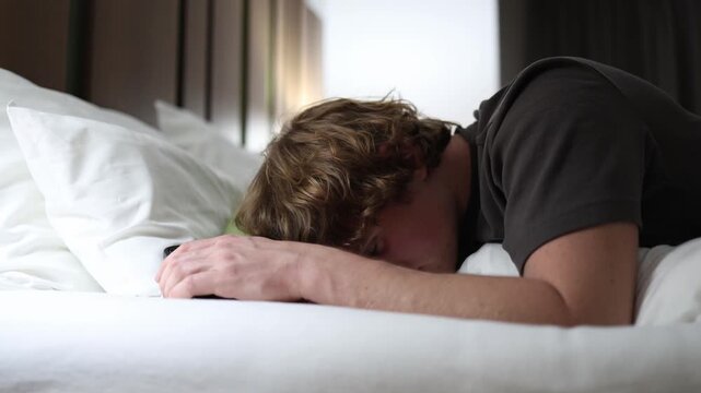 Man lying on bed face down fell asleep holding smartphone exhausted after long scrolling showing digital addiction burnout and insomnia in calm cozy bedroom with soft morning light
