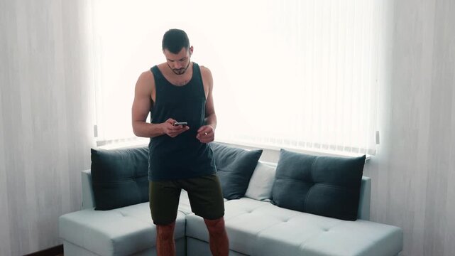 Young man exercising at home. Slow motion of guy turning music on in earphones using smartphone and start running on same spot in room. Warming up body before training and exsercise.