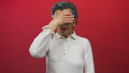 Senior woman with grey hair in white shirt covering her face playfully against a vibrant red...