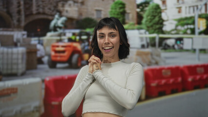 Woman claps hands and smiles in front of building under construction wearing crop top with beaming grin; joy.