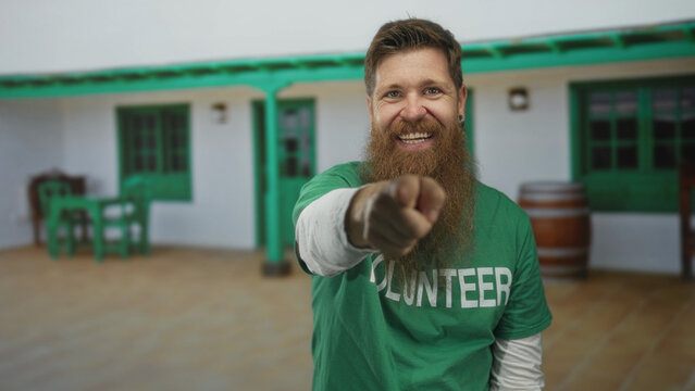 Man wearing green volunteer shirt points finger at camera in front of house entrance; hopeful outreach.