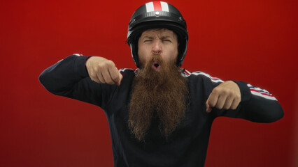 Young bearded man wearing helmet makes motorcycle riding gesture in red studio setting; excitement...