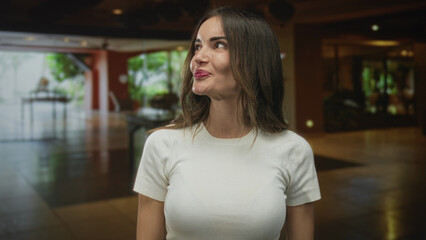 Hispanic woman smiles while pointing both thumbs at herself inside a hotel building under warm lighting; confidence.
