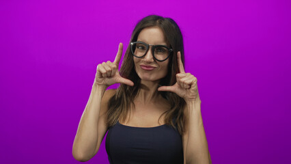 Woman wearing glasses smiles under warm light and uses bare hands to frame her face in a purple studio; creative joy.