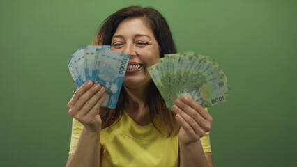 Middle aged hispanic woman fans chilean pesos in a studio with green wall; happiness prosperity...