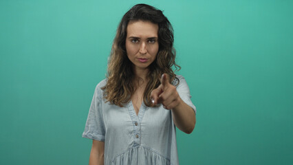 Fototapeta premium Hispanic woman pointing finger at camera and flicking hand to the side in a teal studio; assertion confidence.