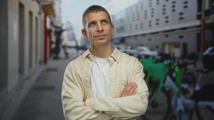 Fototapeta premium Man standing confidently on an urban street with arms crossed, wearing casual beige shirt amidst city environment, showcasing relaxed outdoor vibe in daylight.