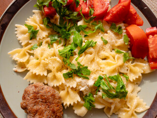 Italian farfalle pasta on a light green plate with grated parmesan, fresh parsley, tomato wedges and a savory meatball for a colorful homemade Italian meal
