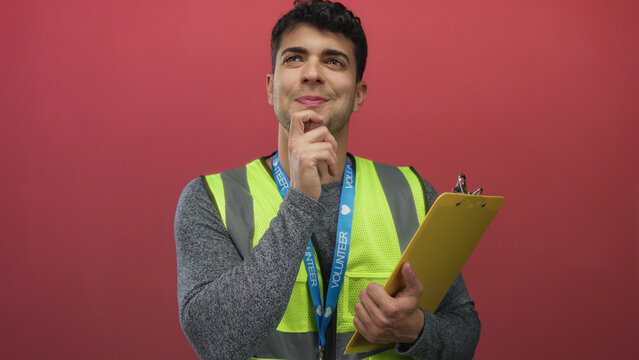 Man touches chin and seems to think against background colored wall while young hispanic volunteer grips clipboard beside guy.